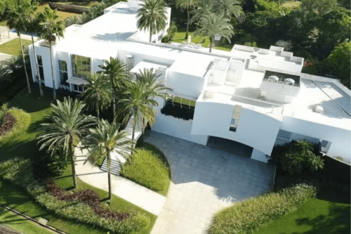 Vista aérea de uma mansão luxuosa de arquitetura contemporânea e minimalista, totalmente pintada de branco. A propriedade possui múltiplos volumes geométricos, telhados planos e grandes janelas de vidro. O paisagismo é composto por gramados impecáveis, diversas palmeiras altas e arbustos ornamentais que cercam uma ampla entrada pavimentada em tons claros.