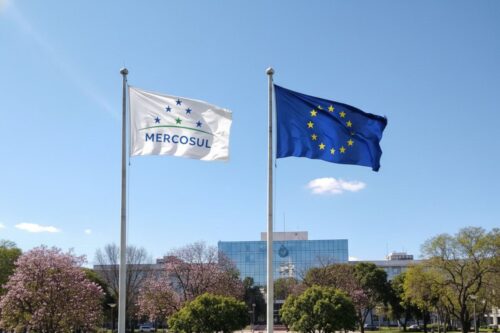 Duas bandeiras hasteadas lado a lado sob um céu azul limpo: à esquerda, a bandeira branca do Mercosul, e à direita, a bandeira azul com estrelas douradas da União Europeia.