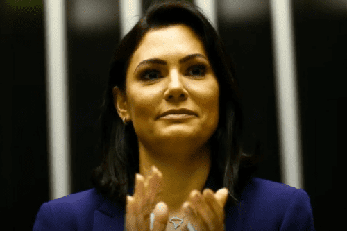 Retrato em plano médio de Michele Bolsonaro, com cabelos escuros na altura dos ombros e vestindo um blazer azul escuro sobre uma blusa branca. Ela aparece aplaudindo, com as mãos em movimento na parte inferior da imagem, e usa um colar com um pingente no formato do mapa do Brasil. O fundo é escuro com linhas verticais claras desfocadas.