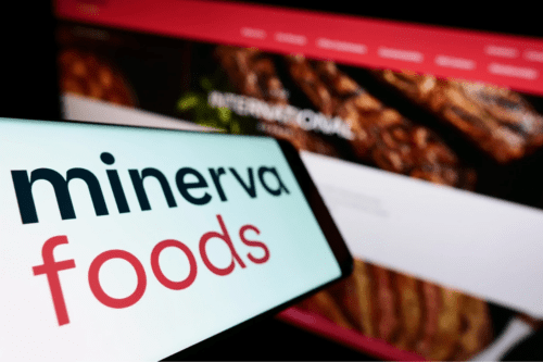 Composição fotográfica focada na marca Minerva Foods. Em primeiro plano, a tela de um smartphone exibe o logotipo da empresa com a palavra "minerva" em preto e "foods" em vermelho sobre fundo branco. Ao fundo, de forma desfocada, aparece a tela de um computador mostrando o site institucional da companhia com imagens de carnes grelhadas.
