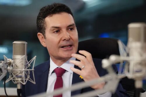 O Ministro das Cidades, Jader Filho, de terno azul e gravata bordô, falando em um microfone de rádio durante uma entrevista em um estúdio.