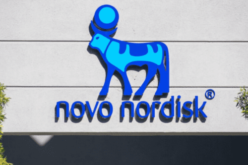 Logotipo da empresa farmacêutica Novo Nordisk fixado em uma parede externa cinza. O logo apresenta a figura estilizada do touro egípcio Ápis em azul, com um círculo acima de seus chifres. Abaixo do símbolo, o nome "novo nordisk" está escrito em letras minúsculas azuis, com o símbolo de marca registrada (R) ao lado.