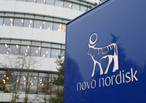 Placa com o logotipo da Novo Nordisk em frente a prédio corporativo moderno, com fachada de vidro e iluminação interna visível.