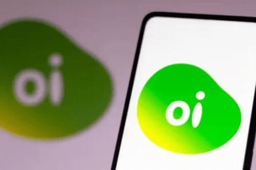 Close em um smartphone exibindo o logotipo da operadora de telefonia Oi na tela. O logotipo consiste na palavra "oi" escrita em letras minúsculas brancas, inserida dentro de uma forma orgânica e fluida com degradê nas cores verde e amarelo. Ao fundo, fora de foco, aparece uma versão maior do mesmo logotipo sobre uma superfície clara.