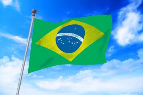 Bandeira do Brasil hasteada em um mastro prateado, ondulando contra um céu azul vibrante com nuvens brancas esparsas.