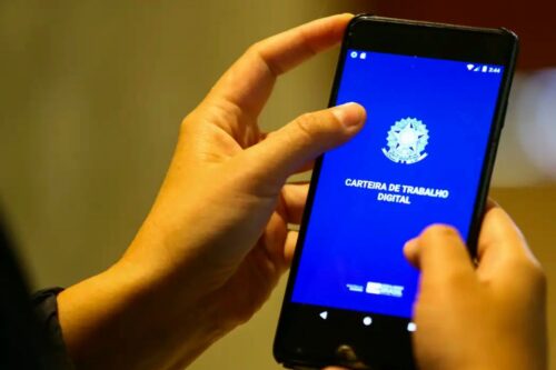 Close-up das mãos de uma pessoa segurando um smartphone que exibe a tela inicial azul do aplicativo Carteira de Trabalho Digital, com o brasão da República e o título em destaque.