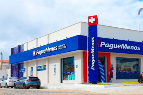 Fachada de uma unidade da rede de Farmácias Pague Menos em uma esquina, destacando o letreiro azul com o logotipo da marca, um totem vertical azul com uma cruz vermelha no topo e carros estacionados à frente sob um céu claro.