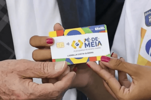 Duas mãos seguram o cartão oficial do programa "Pé-de-Meia: A poupança do ensino médio". O cartão é colorido com as cores da bandeira do Brasil