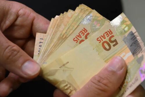 Close das mãos de uma pessoa contando um maço de notas de 50 reais brasileiros, com foco na textura do papel-moeda.