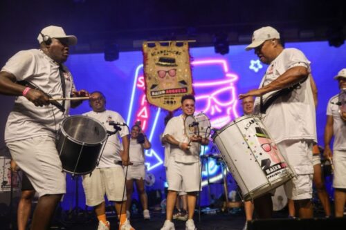 Integrantes da bateria do bloco Acadêmicos do Baixo Augusta tocando instrumentos de percussão no palco durante uma apresentação noturna com luzes neon ao fundo.