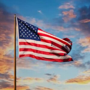 A bandeira dos Estados Unidos hasteada e ondulando ao vento sob um céu dinâmico. O fundo mostra nuvens dispersas iluminadas pelos tons quentes de um pôr do sol dourado e azul.