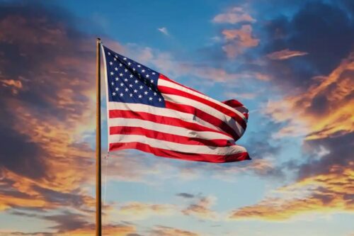 A bandeira dos Estados Unidos hasteada e ondulando ao vento sob um céu dinâmico. O fundo mostra nuvens dispersas iluminadas pelos tons quentes de um pôr do sol dourado e azul.