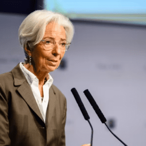 Fotografia de meio corpo de Christine Lagarde, presidente do Banco Central Europeu, falando ao microfone em um púlpito. Ela tem cabelos brancos curtos, usa óculos de armação transparente, brincos de pingente e veste um blazer marrom sobre uma camisa branca. O fundo está desfocado, mostrando tons de azul e cinza.