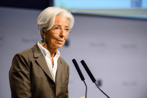 Fotografia de meio corpo de Christine Lagarde, presidente do Banco Central Europeu, falando ao microfone em um púlpito. Ela tem cabelos brancos curtos, usa óculos de armação transparente, brincos de pingente e veste um blazer marrom sobre uma camisa branca. O fundo está desfocado, mostrando tons de azul e cinza.