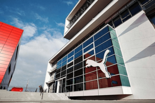 Fachada moderna da sede da Puma, apresentando um edifício de arquitetura contemporânea com grandes painéis de vidro. Em destaque, o icônico logotipo do "jumping cat" da Puma em branco está aplicado sobre a vidraça frontal. Ao lado, parte de uma estrutura vermelha vibrante contrasta com o céu azul e poucas nuvens.