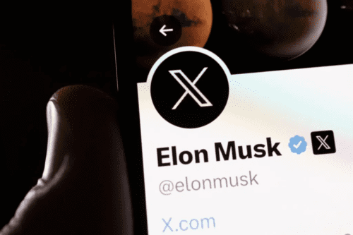 Close do topo da tela de um smartphone exibindo o perfil oficial de Elon Musk na rede social X. O logo da plataforma (um 'X' estilizado em branco sobre fundo preto) está em destaque ao lado do nome 'Elon Musk', que possui um selo de verificado azul. No topo da tela, vê-se parte de uma imagem de capa com o planeta Marte.