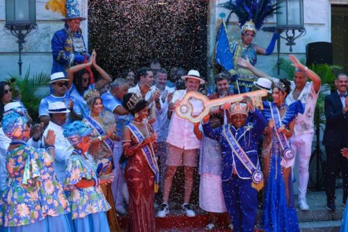 Registro festivo da cerimônia de entrega da chave da cidade no Rio de Janeiro. No centro, o Rei Momo, vestindo um terno azul brilhante e coroa, segura uma grande chave dourada simbólica junto a outras pessoas.
