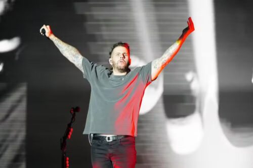Um cantor se apresentando em um palco iluminado. Ele está com os braços abertos e levantados, vestindo uma camiseta cinza básica e calça preta.