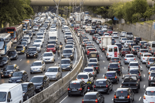 Vista aérea de uma avenida movimentada em São Paulo com tráfego intenso de veículos em diversas faixas. Carros de diferentes modelos e cores, ônibus e algumas motocicletas preenchem a via, sob um viaduto ao fundo e árvores nas laterais.