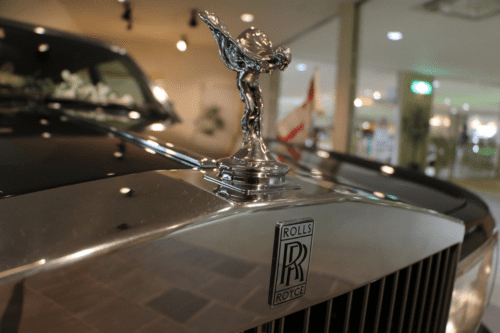 Close-up da parte frontal de um carro de luxo Rolls-Royce preto. Em destaque, sobre a grade cromada, está a icônica estatueta prateada "Spirit of Ecstasy" (Espírito do Êxtase), que representa uma mulher inclinada para frente com os braços estendidos para trás. Logo abaixo, o emblema retangular com o logotipo "RR" da marca está gravado no metal polido.