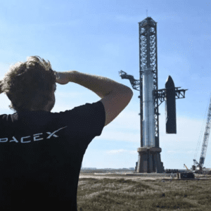 Um homem de costas, vestindo uma camiseta preta com o logo da SpaceX, observa um foguete Starship montado em sua torre de lançamento sob um céu claro. Ao lado, vê-se um guindaste de grande porte.