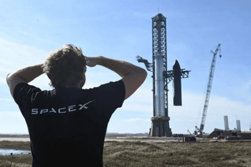 Um homem de costas, vestindo uma camiseta preta com o logo da SpaceX, observa um foguete Starship montado em sua torre de lançamento sob um céu claro. Ao lado, vê-se um guindaste de grande porte.
