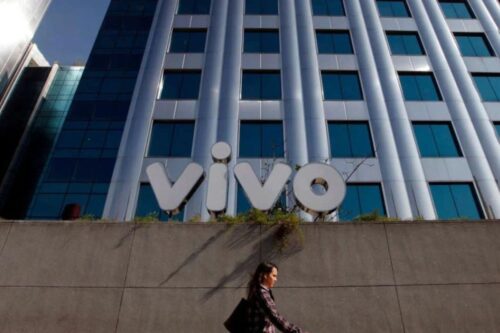 Uma mulher caminha pela calçada em frente à fachada moderna de um edifício corporativo da Telefônica Brasil. No
