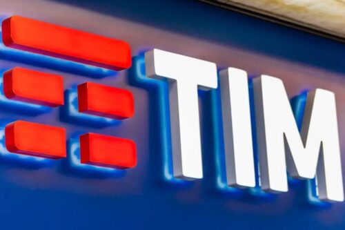 Logotipo tridimensional da operadora TIM instalado em uma fachada. As três barras horizontais vermelhas à esquerda e a palavra 'TIM' em letras brancas se destacam contra um fundo azul iluminado.