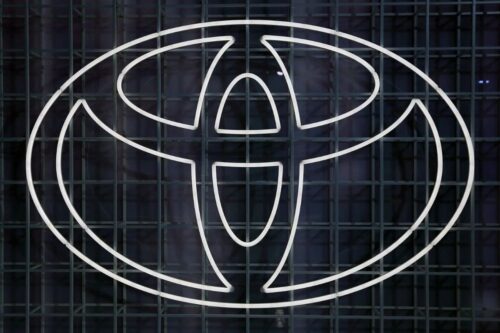 Logotipo da Toyota em formato de letreiro de neon branco brilhante sobre uma grade metálica escura e quadriculada.