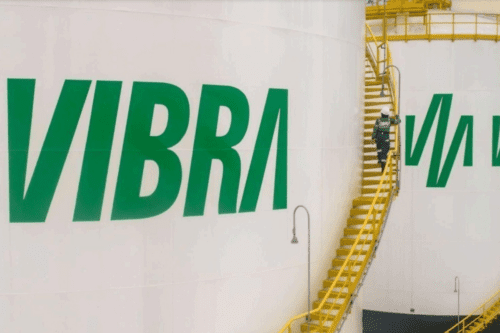 Vista externa de grandes tanques de armazenamento de combustível brancos da empresa Vibra. No tanque em primeiro plano, a palavra "VIBRA" está escrita em letras grandes e verdes. Entre dois tanques, há uma escada metálica amarela externa, por onde sobe um funcionário vestindo uniforme verde e capacete branco de segurança. O céu ao fundo é claro, e a imagem transmite um ambiente industrial organizado.