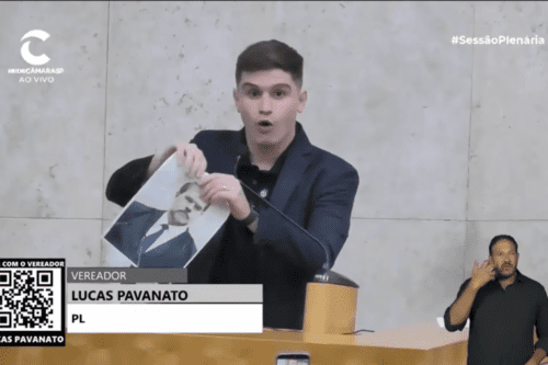 Captura de tela de uma transmissão ao vivo da Rede Câmara SP. No centro, o vereador Lucas Pavanato (PL), um homem jovem de pele clara e cabelos curtos escuros, veste um terno azul-marinho. Ele está atrás de uma tribuna de madeira e segura, com as duas mãos, uma fotografia impressa do ministro Dias Toffoli, iniciando o gesto de rasgá-la ao meio. No canto inferior esquerdo, há um GC (legenda) com seu nome e partido, além de um QR Code. No canto inferior direito, há um intérprete de Libras realizando a tradução sinalizada. O fundo é uma parede de mármore claro.