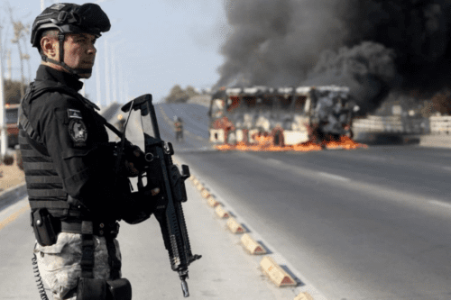 Agente do Ministério Público vigia ônibus incendiado por criminosos em retaliação a operação em Jalisco.