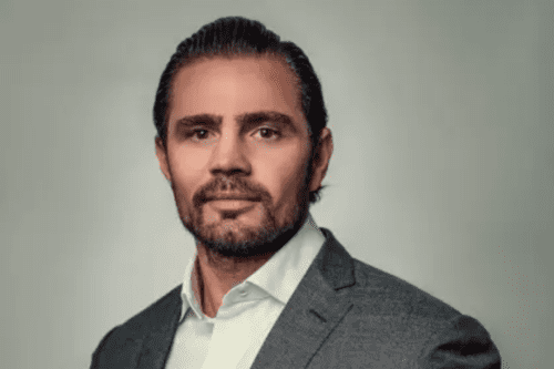 Retrato corporativo de Ricardo Vorcaro. Ele é um homem de pele clara, cabelos escuros penteados para trás e barba bem aparada. Está vestindo um paletó cinza texturizado sobre uma camisa social branca sem gravata. O fundo é neutro em tom cinza claro, transmitindo uma imagem profissional de liderança.