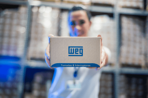 Uma funcionária sorridente, com o rosto levemente desfocado ao fundo, segura e apresenta uma caixa de papelão da empresa WEG em primeiro plano. A caixa exibe o logotipo azul da WEG e o texto "Tomadas & Interruptores", com o ambiente de um estoque ou centro de distribuição visível ao fundo.