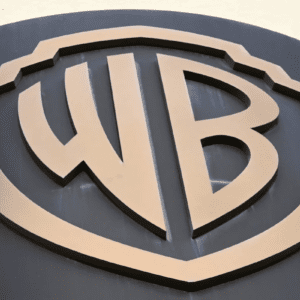 Close-up em ângulo inferior do icônico logotipo de escudo da Warner Bros (WB) em relevo dourado sobre uma superfície escura circular.