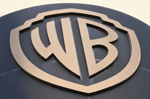 Close-up em ângulo inferior do icônico logotipo de escudo da Warner Bros (WB) em relevo dourado sobre uma superfície escura circular.