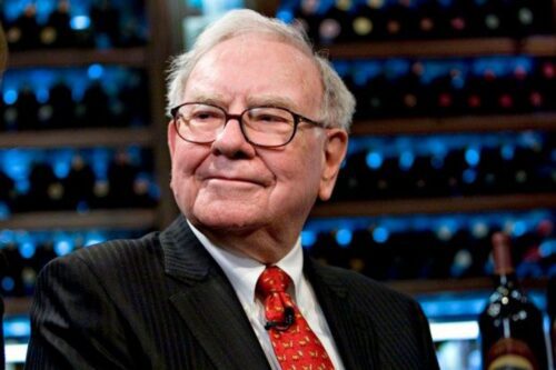Retrato em plano médio do investidor Warren Buffett. Ele sorri levemente, usa óculos de armação escura, terno risca-de-giz preto, camisa branca e uma gravata vermelha com detalhes dourados.