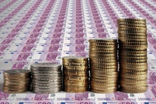 Cinco pilhas de moedas metálicas de tamanhos crescentes, da esquerda para a direita, organizadas como um gráfico de barras sobre um fundo composto por várias notas de 500 euros espalhadas. A imagem sugere crescimento financeiro e acúmulo de capital.