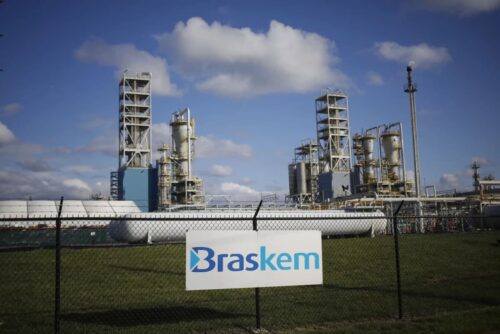 Fachada de unidade industrial da Braskem com estruturas petroquímicas ao fundo e placa da empresa em destaque.