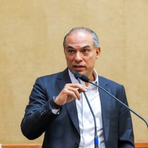 Augusto Ferreira Lima de terno fala ao microfone em tribuna, com bandeira ao fundo em ambiente institucional.