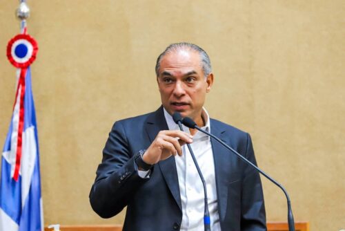 Augusto Ferreira Lima de terno fala ao microfone em tribuna, com bandeira ao fundo em ambiente institucional.