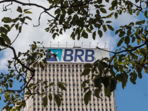 Fachada de prédio corporativo com logotipo do Banco BRB no topo, vista entre galhos e folhas de árvores.