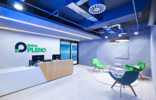 Recepção moderna do Banco Pleno com logo na parede, balcão de atendimento e poltronas verdes em sala iluminada.