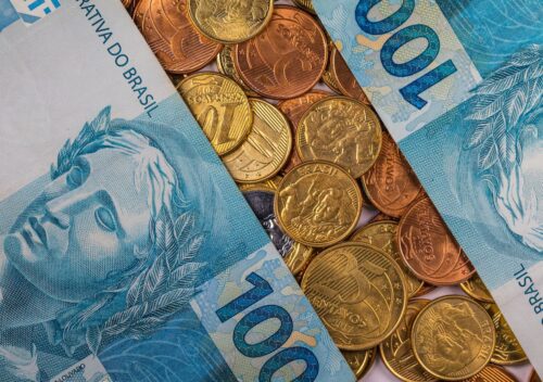 Notas de 100 reais e moedas espalhadas, representando dinheiro e economia no Brasil.