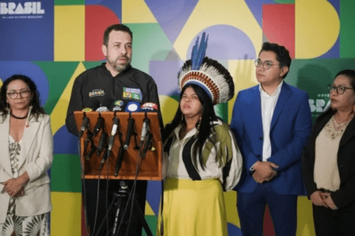 Fotografia de uma coletiva de imprensa com cinco autoridades brasileiras à frente de um painel colorido do Governo Federal. Ao centro, Guilherme Boulos fala ao microfone em um púlpito de madeira repleto de microfones de diversas emissoras. À sua direita, a ministra Sonia Guajajara, usando um cocar tradicional indígena e vestes em tons de amarelo e preto. À esquerda, uma mulher de blazer branco e óculos. À direita de Sonia, um homem de terno azul e óculos, e na extremidade direita, outra mulher de blazer escuro. O ambiente sugere um pronunciamento oficial.