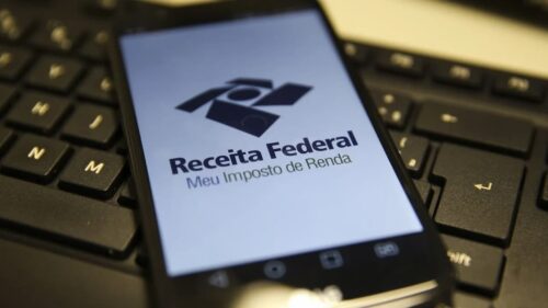 Celular exibe aplicativo Meu Imposto de Renda da Receita Federal sobre teclado de computador.