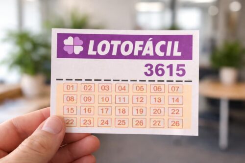 Mão segurando bilhete da Lotofácil 3615 com grade de números, em ambiente interno desfocado ao fundo.