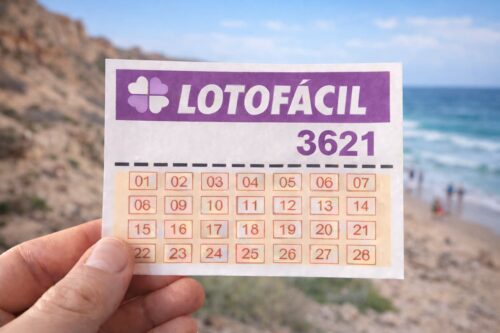 Bilhete Lotofácil 3621 segurado diante de praia com falésias e pessoas ao fundo desfocado.