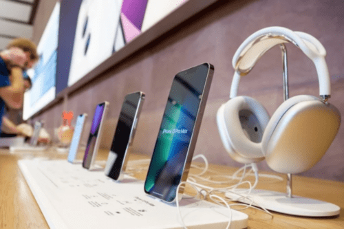 Diversos produtos da Apple em exposição sobre uma bancada de madeira clara em uma loja. Em destaque, um iPhone 13 Pro Max inclinado e um fone de ouvido AirPods Max branco em um suporte. Ao fundo, outros modelos de smartphones e clientes desfocados, ilustrando o mercado de tecnologia e bens de consumo.
