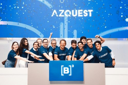 Equipe da AZQuest comemora evento na B3. O grupo aparece sorridente, vestindo camisas personalizadas da empresa, em frente ao painel oficial da Bolsa de Valores.